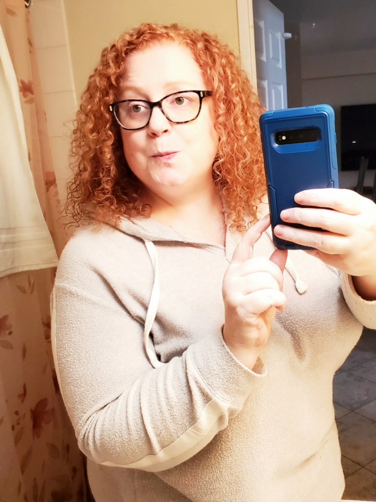 Selfie featuring a red haired woman with curly hair and dark brown glasses named Jenn standing in front of a mirror