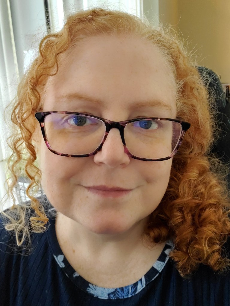 Selfie featuring a red haired woman with curly hair and dark brown glasses named Jenn